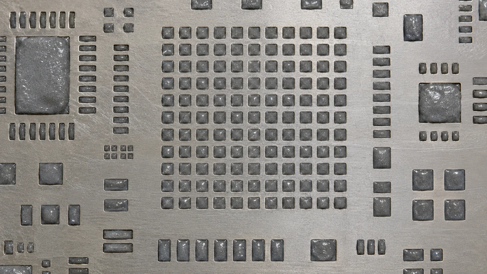 Gray solder paste sitting in the openings of a bare metal stencil.