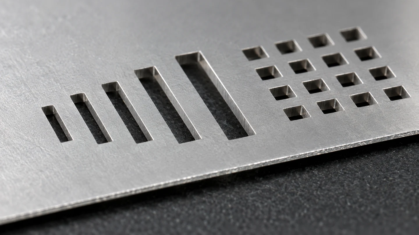 Close view of rectangular and square openings through stainless steel stencil foil, with the sheet edge visible.